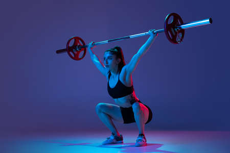 Portrait of muscled woman in sportswear training with a barbell isolated on purple background in neon light. Sport, weightlifting conceptの写真素材
