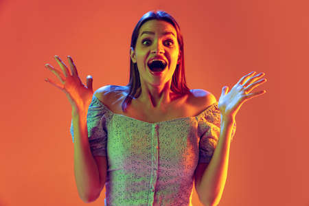 Wow. Half-length emotional young girl with long shiny hair isolated on peach color background in neon filter. Concept of emotions, beauty, fashion, youthの写真素材