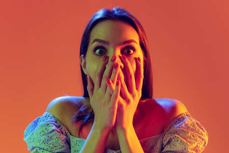 Scared young girl with long shiny hair isolated on peach color background in neon filter. Concept of...
