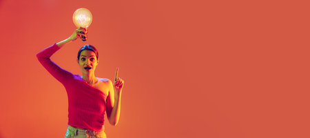 Flyer with emotional young girl holding big electric bulb over her head isolated on peach color background in neon filter. Concept of emotions, beauty, fashion, youthの写真素材