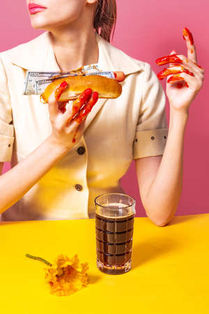Food pop art photography. Young woman tasting hot dog with dark beer isolated on bright pink and yellow background. Vintage, retro style interiorの写真素材