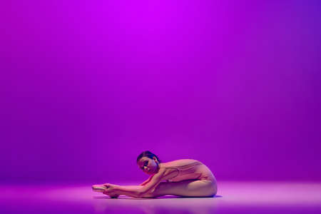 One flexible young female ballet dancer, teen in stage outfit and pointes sitting on floor isolated on purple background in neon light. Art, grace, beauty, ballet school conceptの写真素材