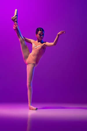 Portrait of young little ballet dancer, teen practicing, dancing isolated on purple background in neon light. Art, grace, beauty, ballet school conceptの写真素材