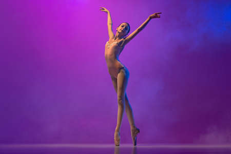 Beautiful flexible young female ballet dancer, teen in stage outfit and pointes dancing isolated on purple background in neon light with smoke. Art, grace, beautyの写真素材