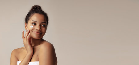 Portrait of young beautiful woman applying moisturizing cream, taking care after skin isolated over grey backgroundの写真素材