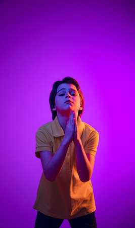 Portrait of emotive boy, child having hands in praying position, posing isolated over purple...