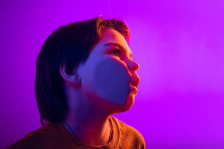 Close-up boy, kid leaning against transparent glass by cheek isolated on purple studio background in...