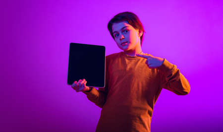 Portrait of boy, child pointing at tablet isolated over purple background in neon light