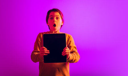 Portrait of boy, child with expressive emtions holding tablet isolated over pink background in neon lightの写真素材