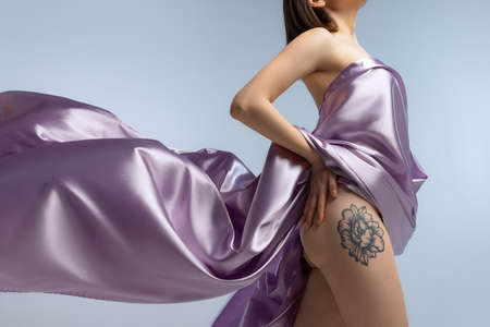 Cropped studio shot of slim female body covered with purple silk cloth isolated over blue studio...