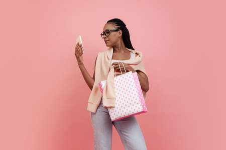 Portrait of young charming girl with afro hairdo in casual style outfit holding shopping bags isolated on pink background. Concept of beauty, art, fashion, youth, styleの写真素材
