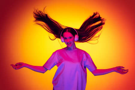 Emotional caucasian girl wearingt-shirt dancing, listening to music isolated on yellow-red background in neon. Concept of beauty, art, fashion, youth and emotionsの写真素材