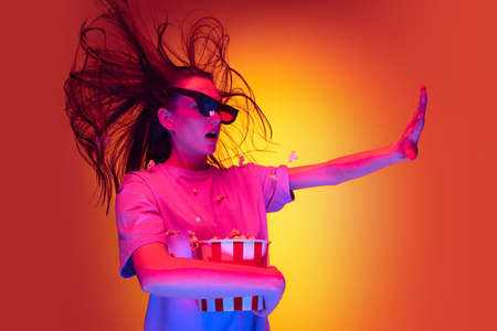 One excited young girl, student wearing 3d glasses and holding bucket of popcorn isolated on orange...
