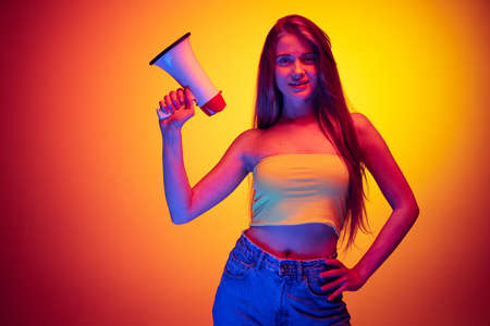 Portrait of young beautiful girl posing with megaphone isolated over gradient red yellow background in neon lightの写真素材