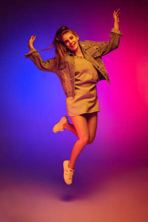 Portrait of young girl in casual cloth jumping, posing isolated over gradient pink blue background...