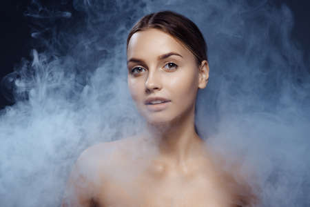 Portrait of beautiful young girl with clean, well kept skin isolated on dark background with smoke. Concept of beauty, art, ad, cosmeticsの写真素材