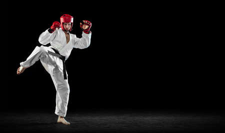 Male taekwondo fighter in white dobok, helmet and gloves training isolated over dark background. Concept of sport, workout, competition, adの写真素材