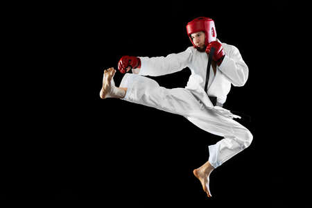 Male taekwondo fighter in white dobok, helmet and gloves training isolated over dark background....
