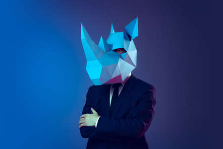 Creative portrait of young stylish man in business suit with cardboard mask on his head isolated on dark blue studio background. Concept of art, fashion, theaterの写真素材