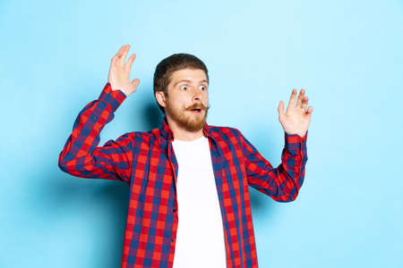 Emotional young red-headed man in white t-shirt and plaid shirt posing isolated on blue background....