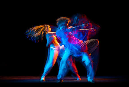 Excited man and woman dancing hip-hop in sportive style clothes on dark background at dance hall in mixed neon light. Youth culture, hip-hop, movementの写真素材