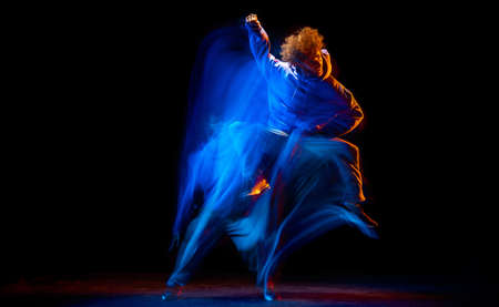 Young sportive man dancing hip-hop in sportive style clothes isolated on dark background at dance hall in mixed neon light. Youth culture, hip-hop, movementの写真素材