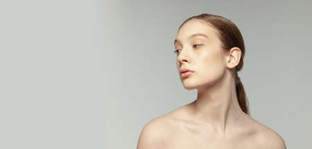 Tender young girl with low ponytail and no makeup face posing isolated over grey studio background....