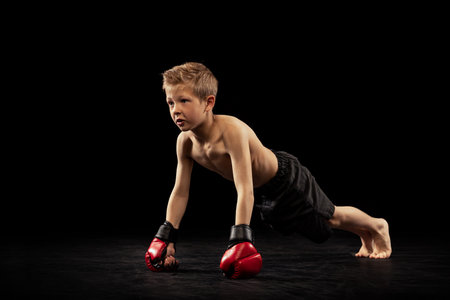 Studio shot of preschool boy, cute kid in sports shots and gloves training alone on black...
