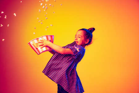 One happy little girl, pupil wearing 3d glasses and holding bucket of popcorn isolated on orange background in neon. Concept of emotions, facial expression, youth, aspirationの写真素材