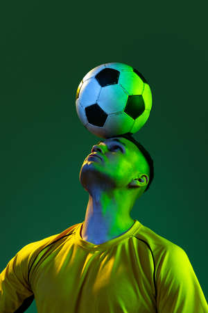 Half-length of professional male football soccer player playing with ball isolated on green background in neon light. Concept of sport, goals, competition, game, achievementsの写真素材