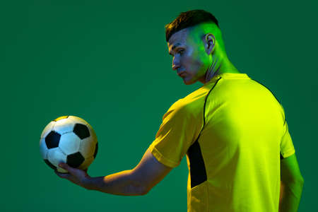 Half-length of professional male football soccer player holding ball isolated on green background in neon light. Concept of sport, goals, competition, game, achievementsの写真素材