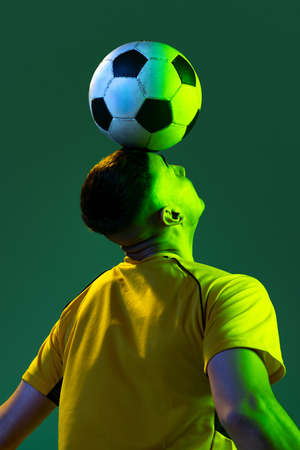 Half-length of professional male football soccer player playing with ball isolated on green background in neon light. Concept of sport, goals, competition, game, achievementsの写真素材