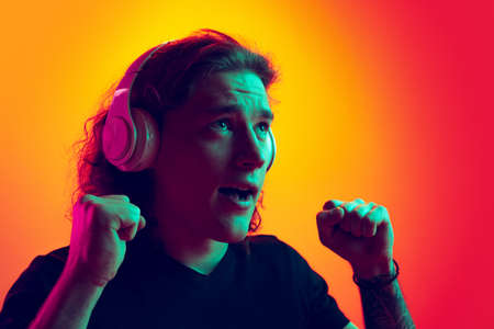 Young excited man with long curly hair listening to music in headhones isolated on gradient yellow-pink background in neon. Concept of beauty, fashion, youth culture and emotionsの写真素材