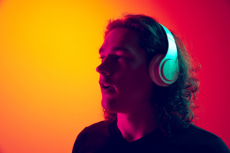 Young excited man with long curly hair listening to music in headhones isolated on gradient...