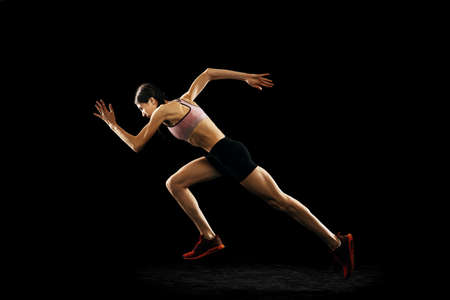 Studio shot of young muscular woman running isolated on black background. Sport, track-and-field athletics, competition and active lifestyle conceptの写真素材