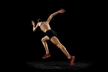 Back view of young muscular woman running isolated on black background. Sport, track-and-field...