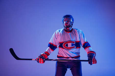 Portrait of professional hockey player in sports uniform and protective equipment isolated on purple background in neon. Concept of sport, healthy lifestyle, hobby, movement, action.の写真素材