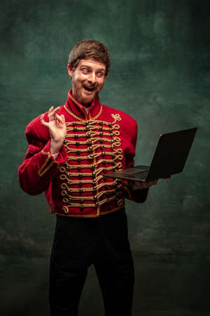 Handsome young man in image of medieval hussar using laptop isolated on dark blue background. Retro...