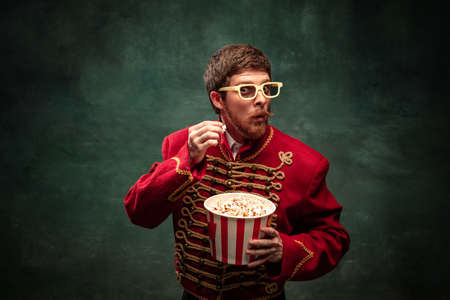 Shocked handsome young man in image of medieval hussar eating popcorn isolated on dark blue...