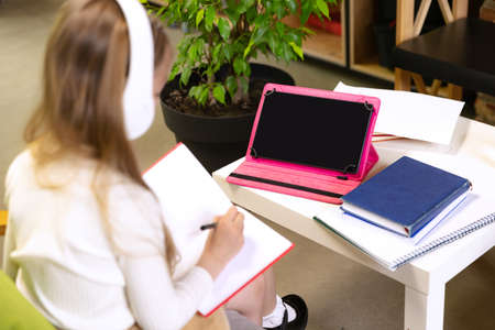 One beautiful little girl, school age kid studying at home using tablet and headphones. Model...