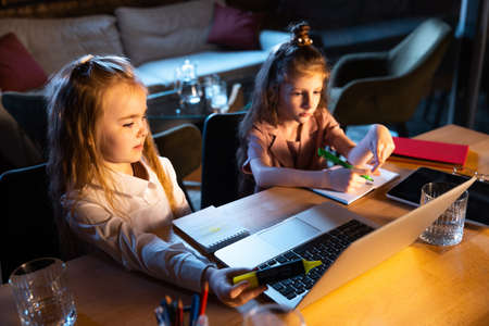 Two little cute kids, sisters sitting at home and doing homework at home interior at evening time....
