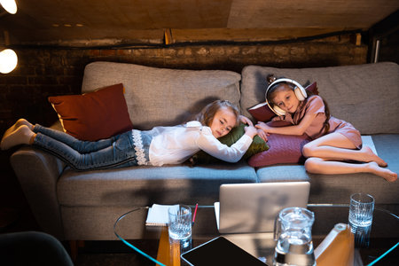 Cute little girls, kids having fun, watching cartoons on laptop at home at evening time. Education, childhood, family, remote learning and emotions concept.の写真素材