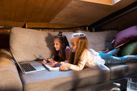 Happy cute little girls, kids having fun, watching cartoons on laptop at home at evening time. Education, childhood, family, remote learning and emotions concept.の写真素材