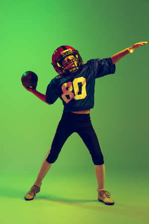 Sportive little boy in sports uniform and equipment playing american football isolated on green...