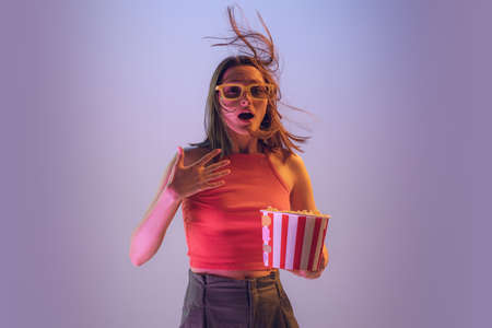 Young shocked woman in casual outfil eating popcorn and watching movie isolated on blue background. Concept of emotions, facial expression, fashion and beautyの写真素材