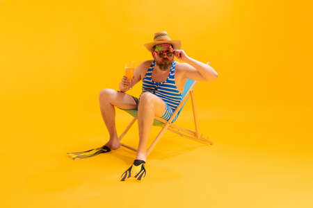 Studio shoot of cool funny beachgoer, man in vintage striped swimsuit and bowler hat sitting in...