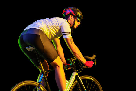 Portrait of young male cyclist on bicycle in cycling shorts and protective helmet isolated on dark background in neon. Concept of sport, speed, energyの写真素材