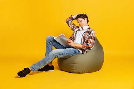 Emotive boy, student in casual style clothes sitting at chair and reading book isolated over yellow background. Back to school, universityの写真素材