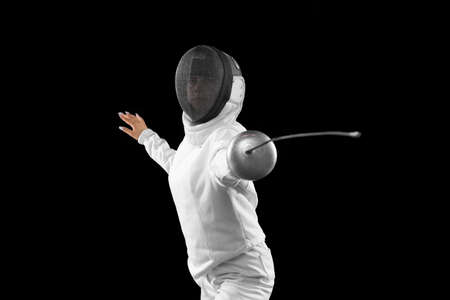 Energetic female fencer in white fencing costume and mask in action, motion isolated on dark background. Sport, youth, activity, skills, achievements, goal.の写真素材