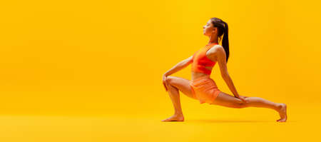 Portrait of young slim, flexible girl in shorts doing stretching exercises isolated on bright yellow...